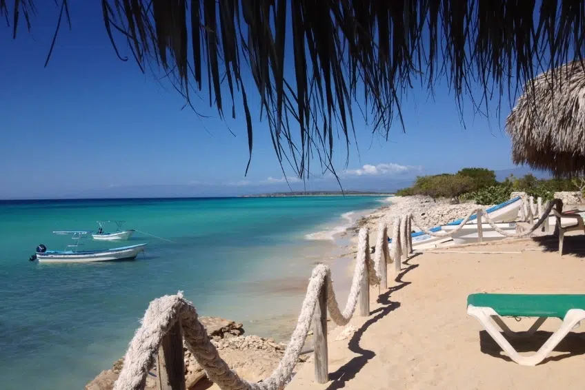 Beach loungers under thatched palapa with boat anchored offshore at Bahia de las Aguilas